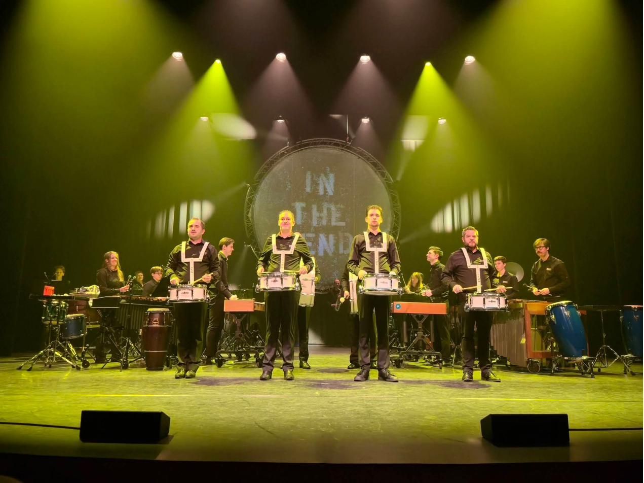 Vier slagwerkers met marstrommels vooraan op het podium, in geel en groen licht
